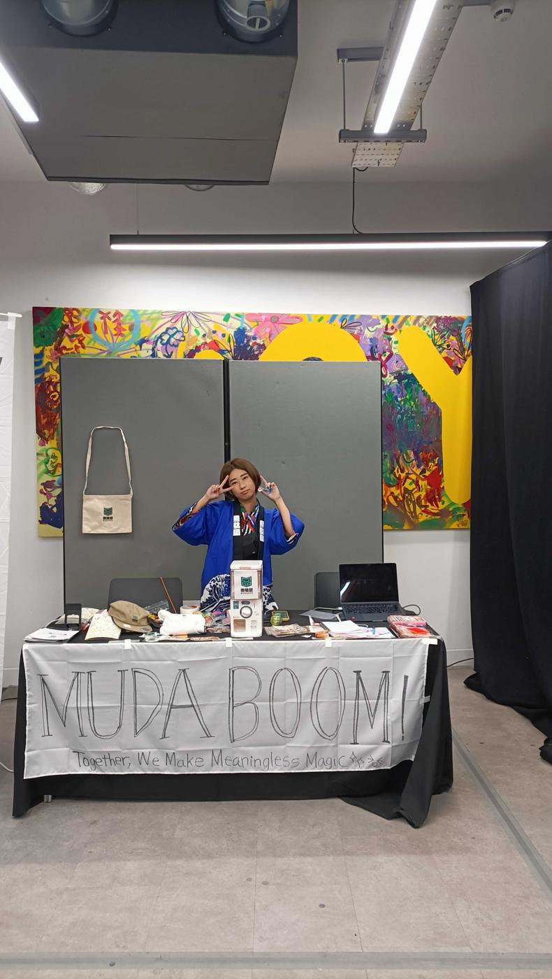 MON-chan behind her booth under a hand-painted banner: MUDA BOOM! Together, we make meaningless magic.