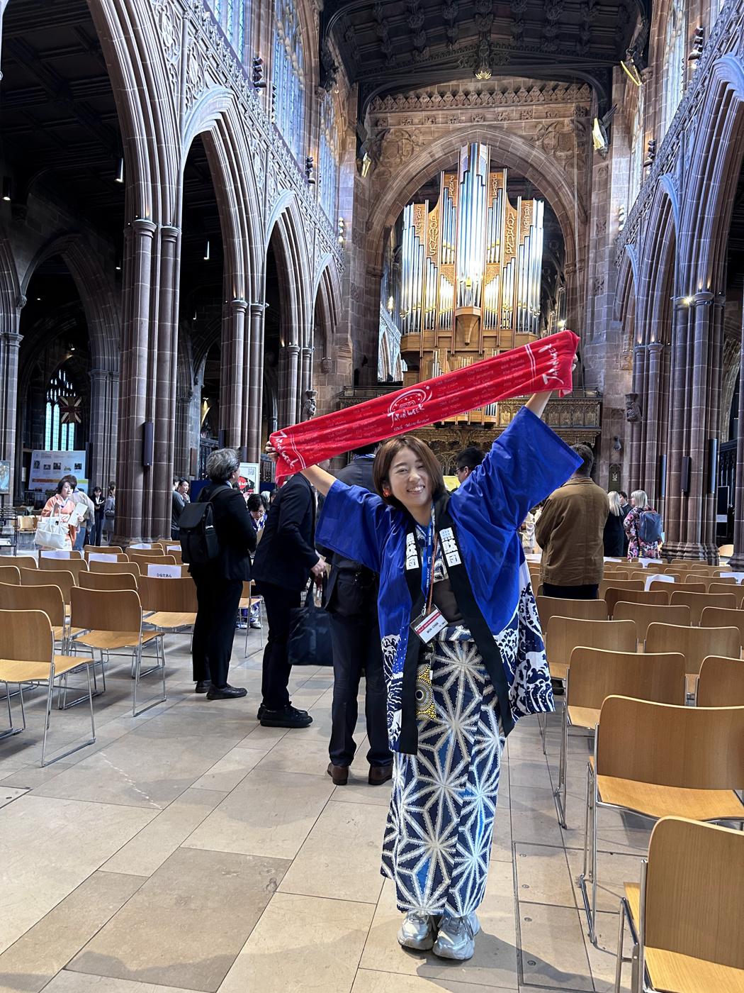 MON-chan in a happi coat holding up a red Japan Week towel inside Manchester Cathedral.