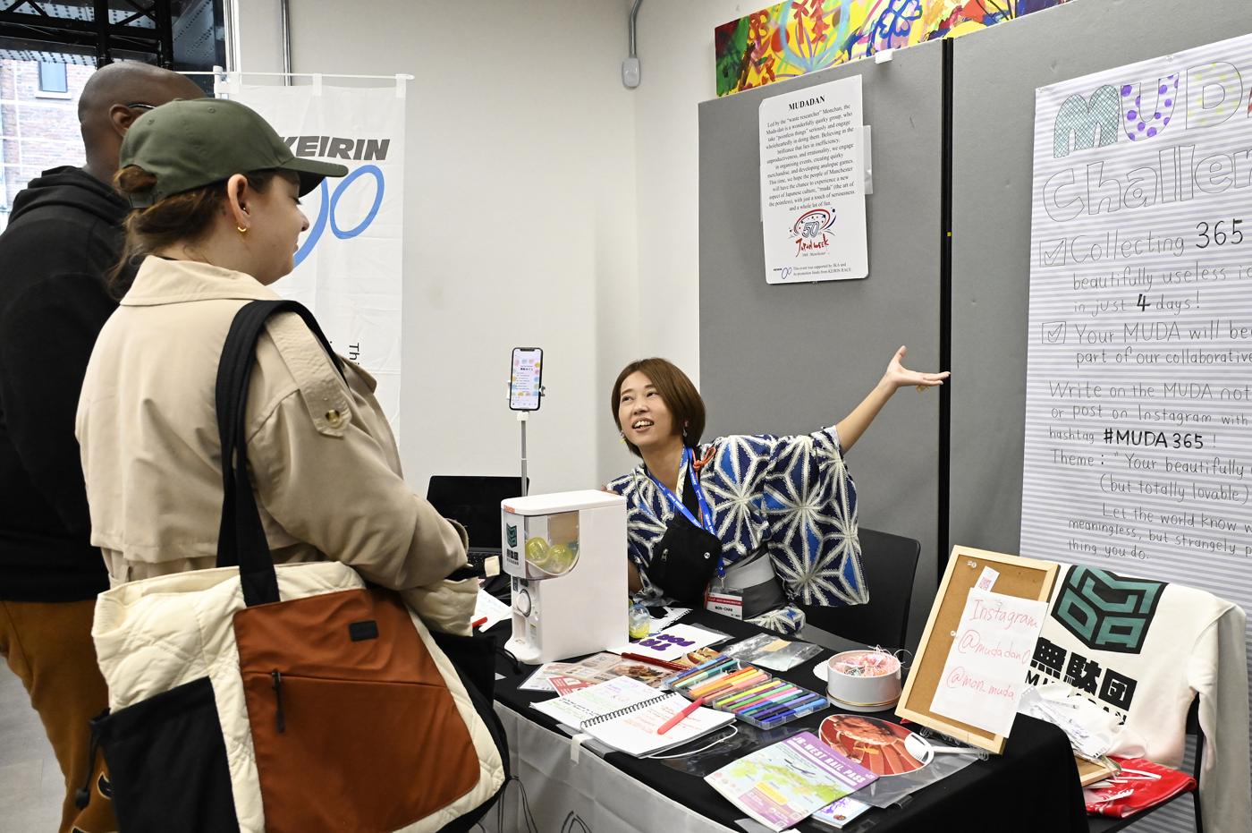 MON-chan at her booth gesturing to a hand-written poster for the MUDA Challenge.
