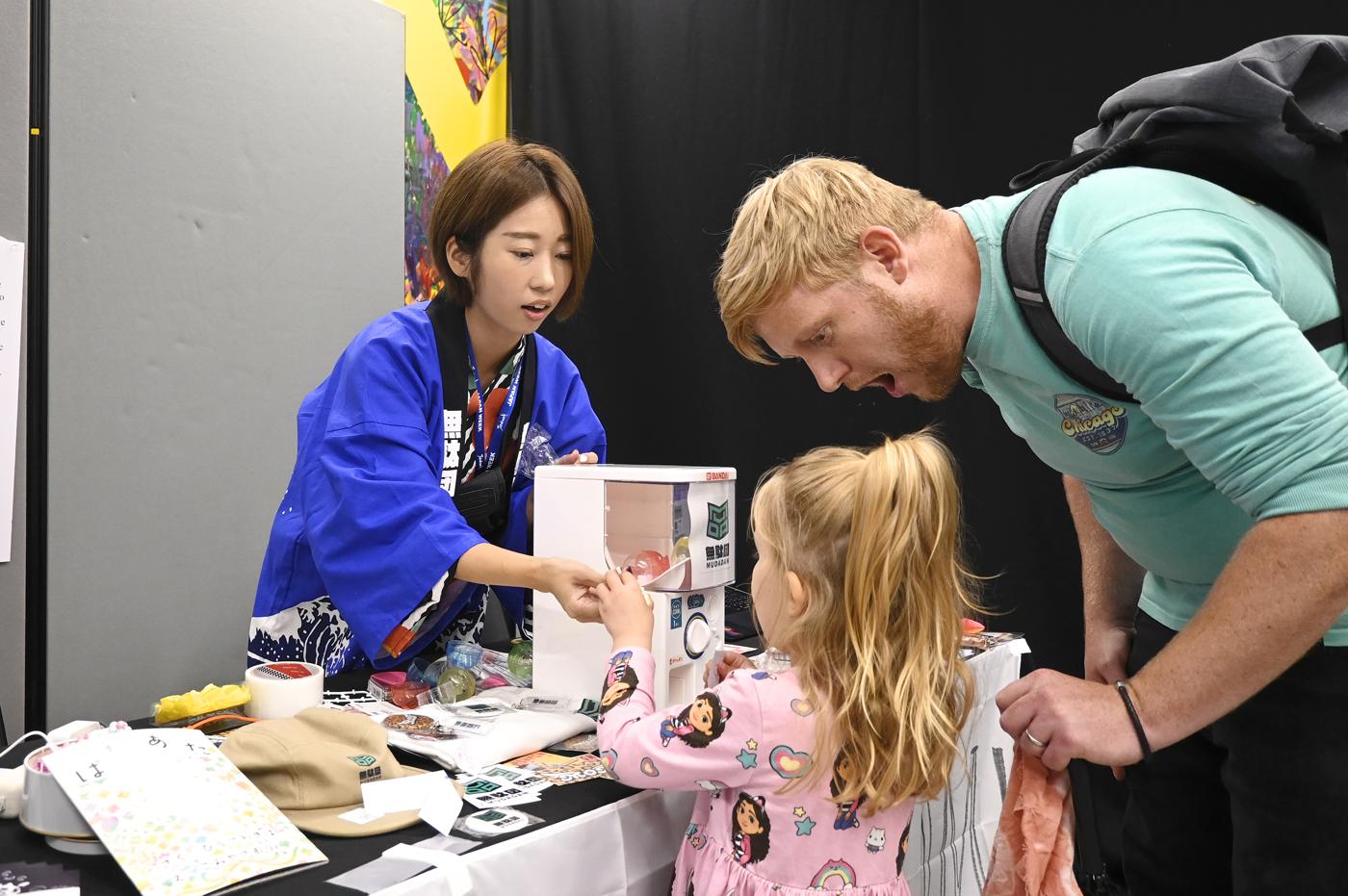 A father gasps with delight as his daughter gets a capsule from the MUDADAN gachapon machine, while MON-chan in a blue happi coat hands it over.