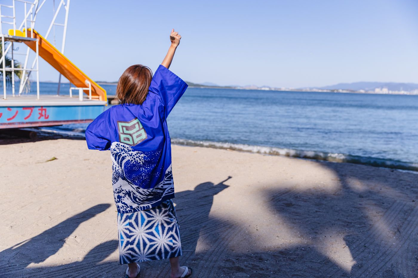MON-chan on a beach in a MUDADAN happi coat, fist raised to the sea