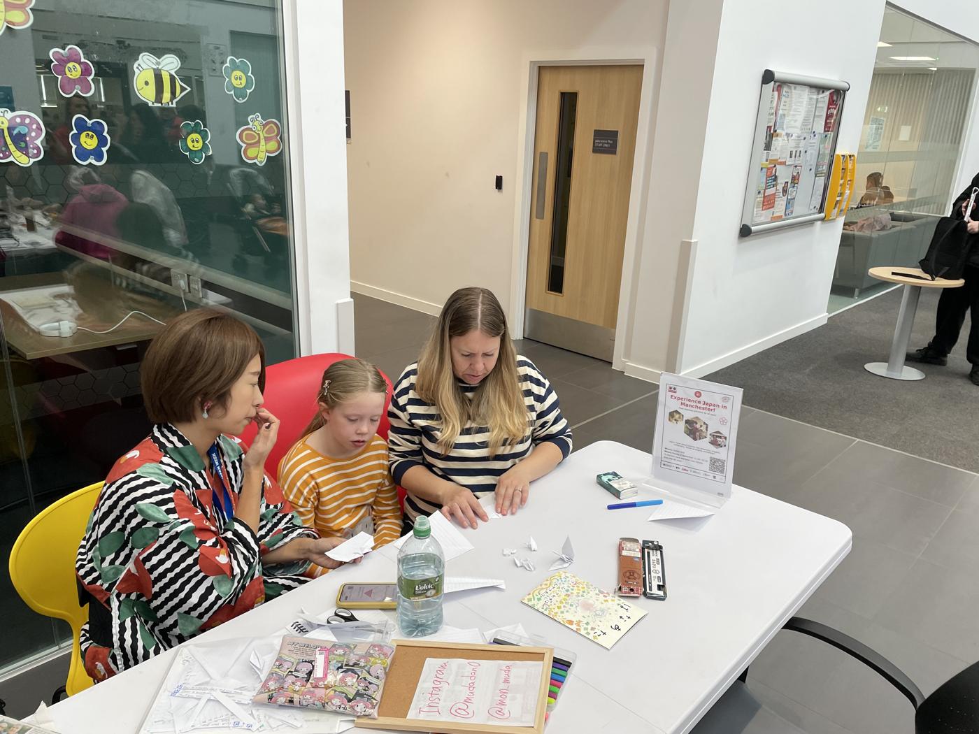 MON-chan folding paper cranes with a girl and her mother at Gorton Library.