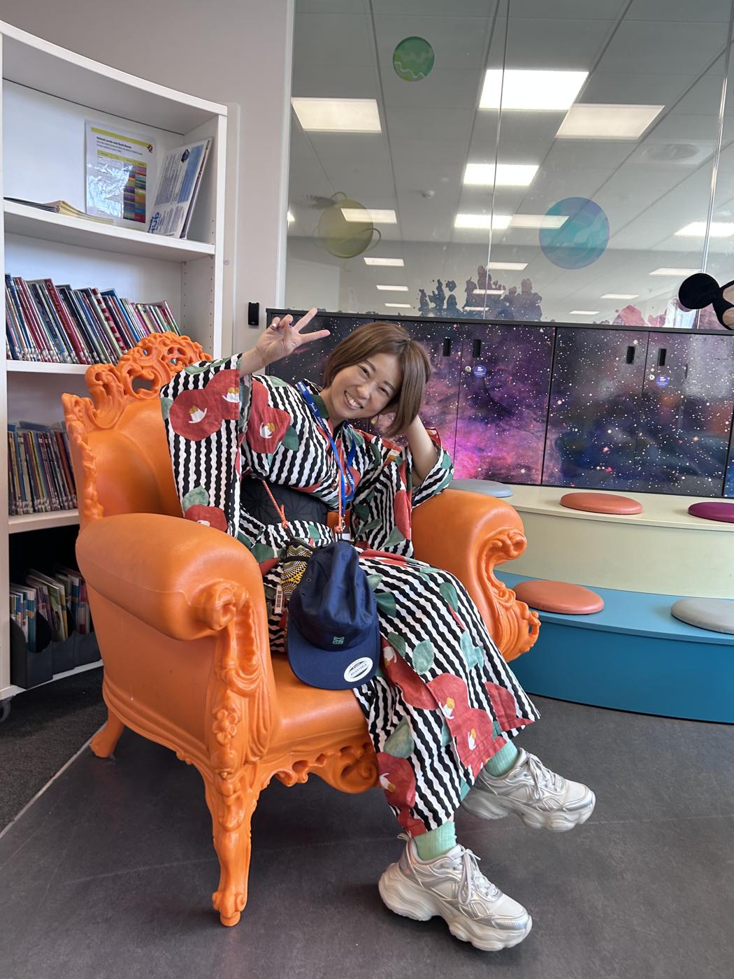 MON-chan sitting sideways in an ornate orange armchair, wearing a striped camellia kimono and sneakers, making a peace sign.